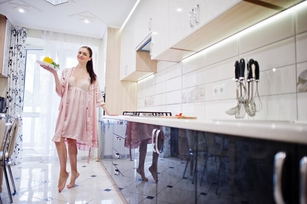 A woman in a pink robe stands in a kitchen, surrounded by cabinets and appliances, looking relaxed and at home